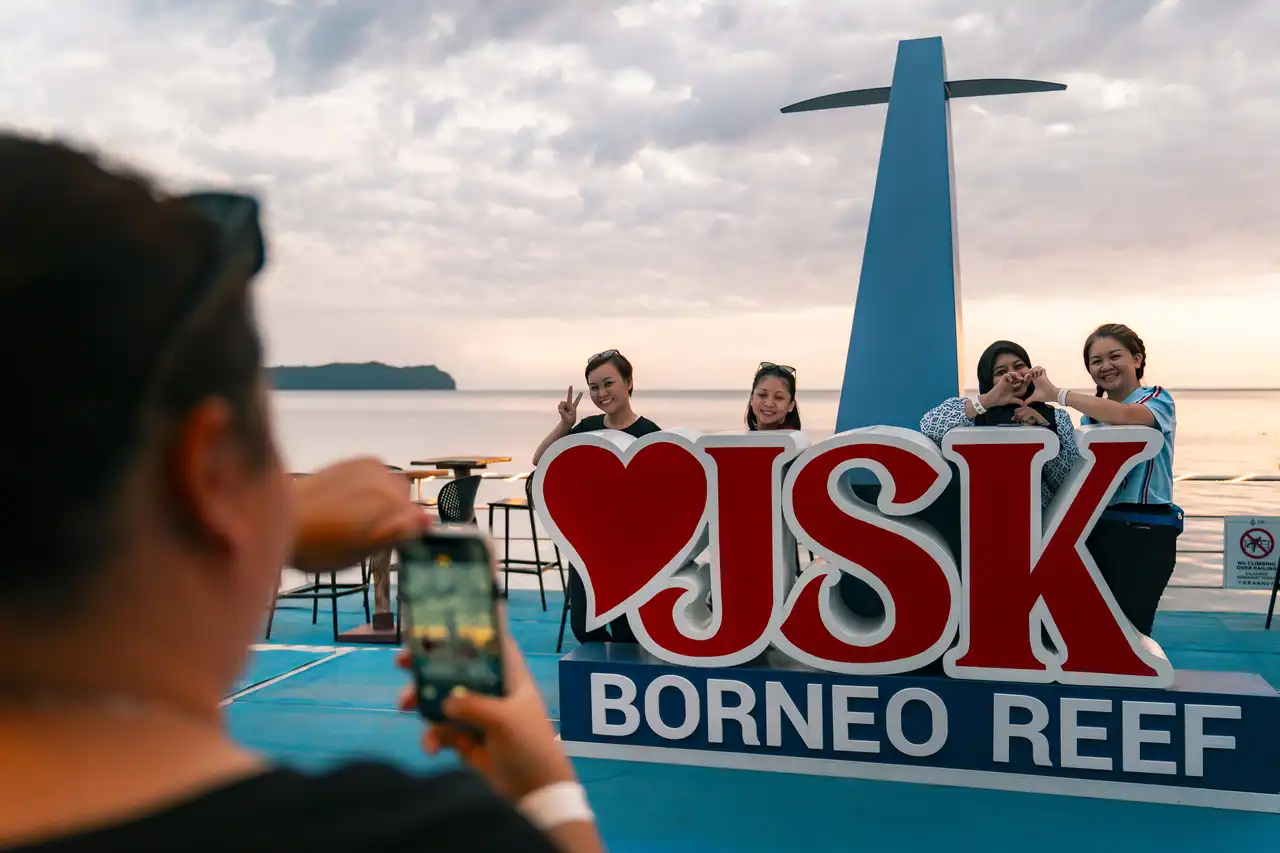 A group of women posing for a photo at the JSK Borneo Reef sign with a scenic ocean backdrop during sunset, while another person takes their photo.