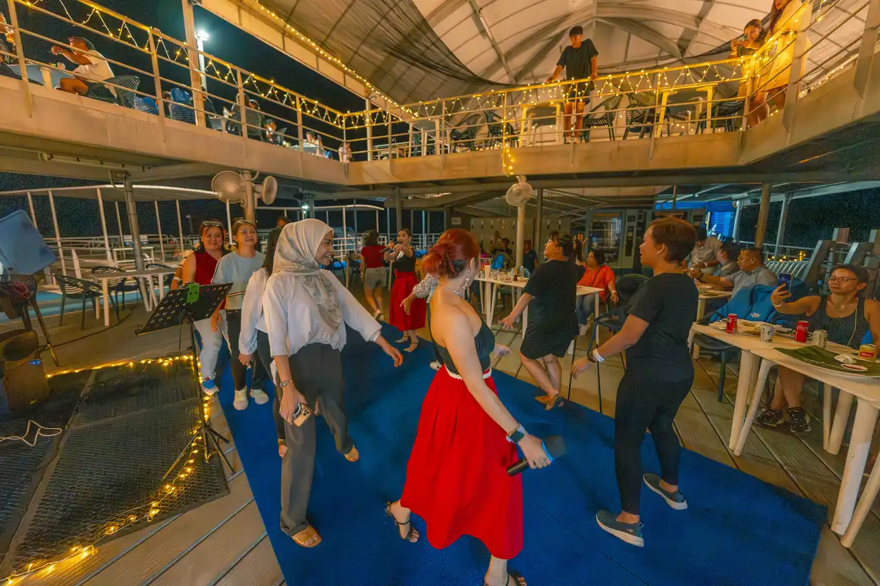A diverse group of people enjoying a lively dance party on a boat deck at night, with some participants dancing energetically while others watch from the upper deck, illuminated by string lights and a warm, festive atmosphere.