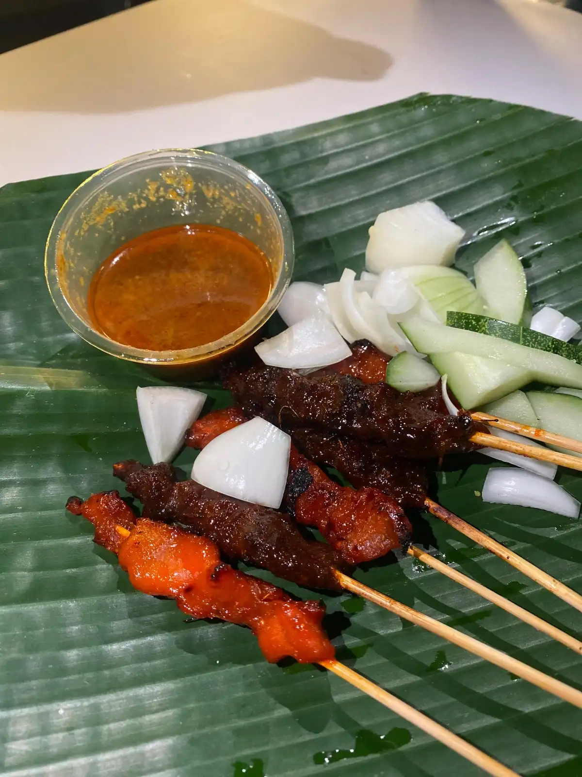 Grilled satay skewers with peanut sauce, cucumber, and onion slices on a banana leaf.
