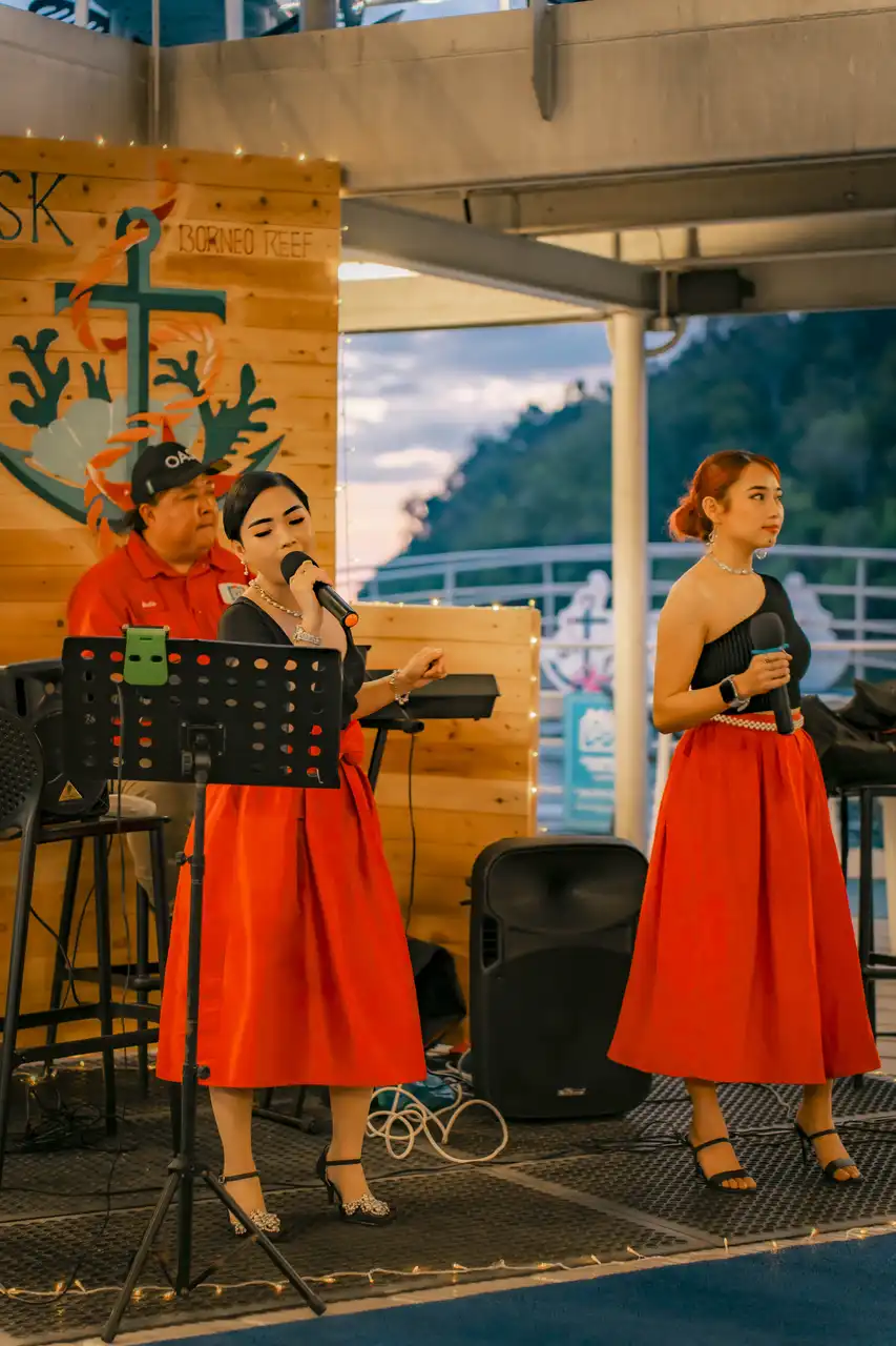 Two women in red skirts performing at a Borneo Reef event, one singing and the other holding a microphone.