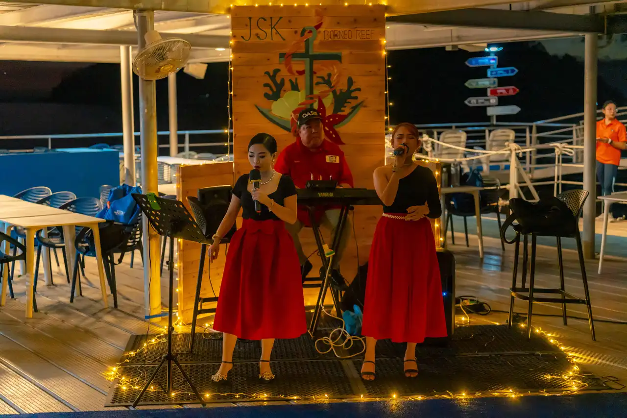 Two women in red dresses singing with microphones and a man playing keyboard on a decorated boat deck at night.