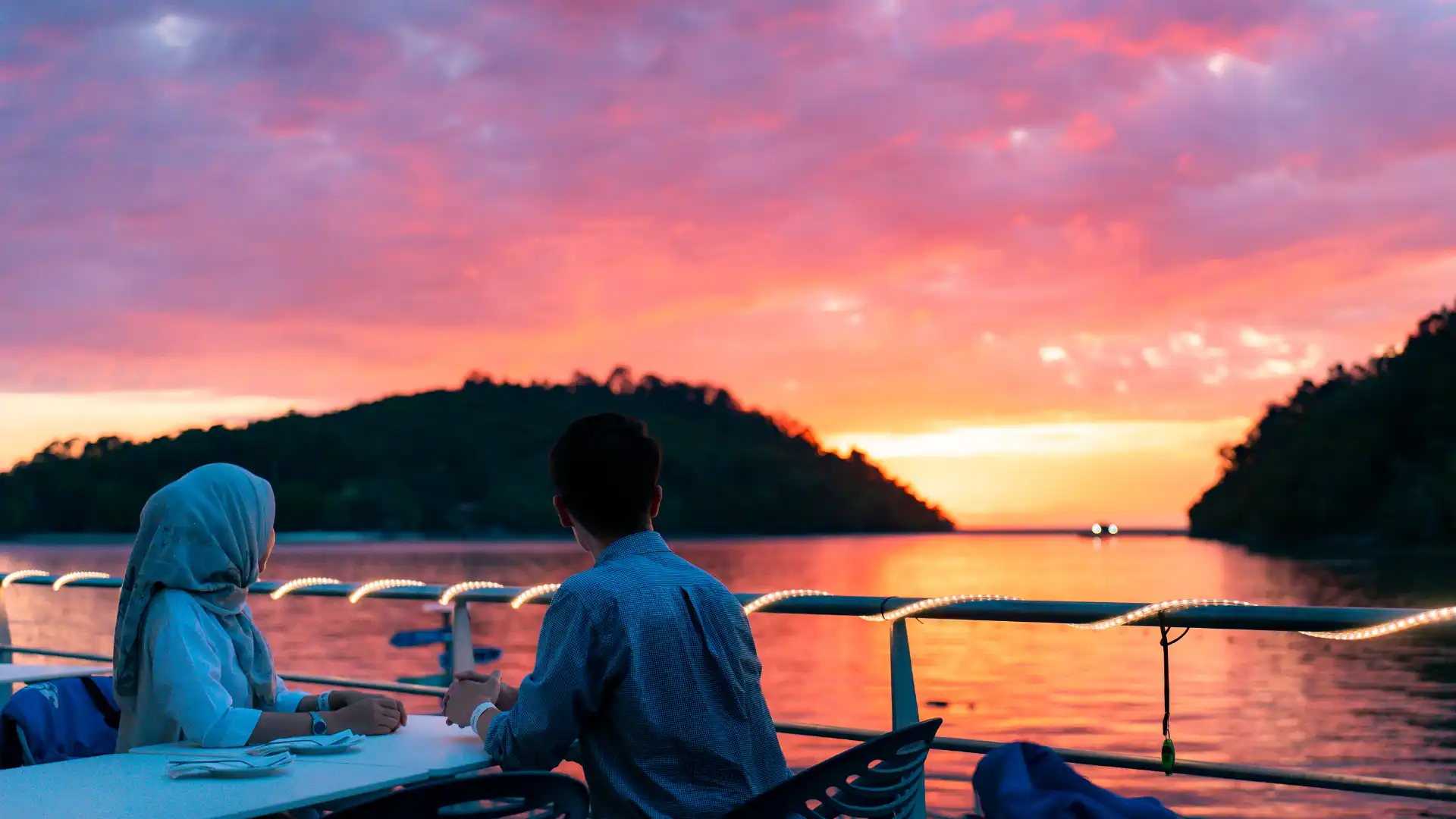 A couple enjoys a sunset dinner on a boat overlooking a scenic lake.