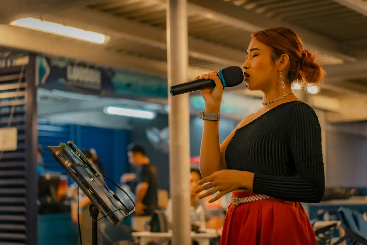 Woman singing into a microphone at a night market with a music stand in the foreground.