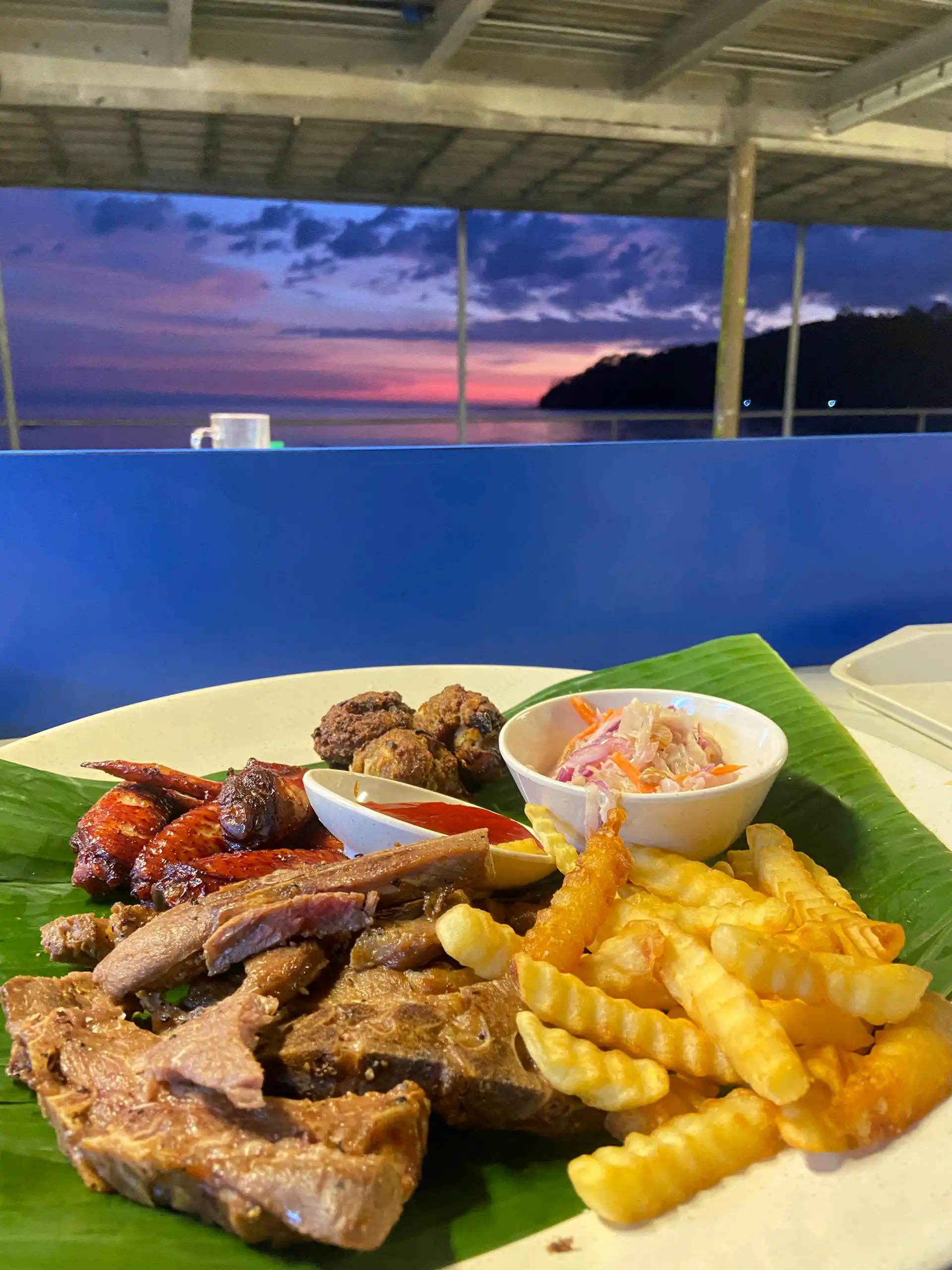 A plate of assorted grilled meats, fries, and coleslaw on a banana leaf, with a sunset over the ocean in the background.