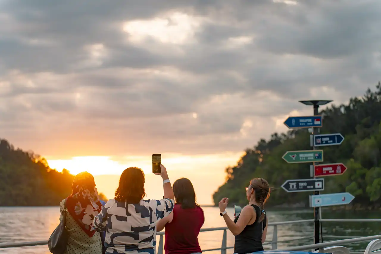 Group of people capturing sunset on their phones by a directional signpost showing distances to global cities.