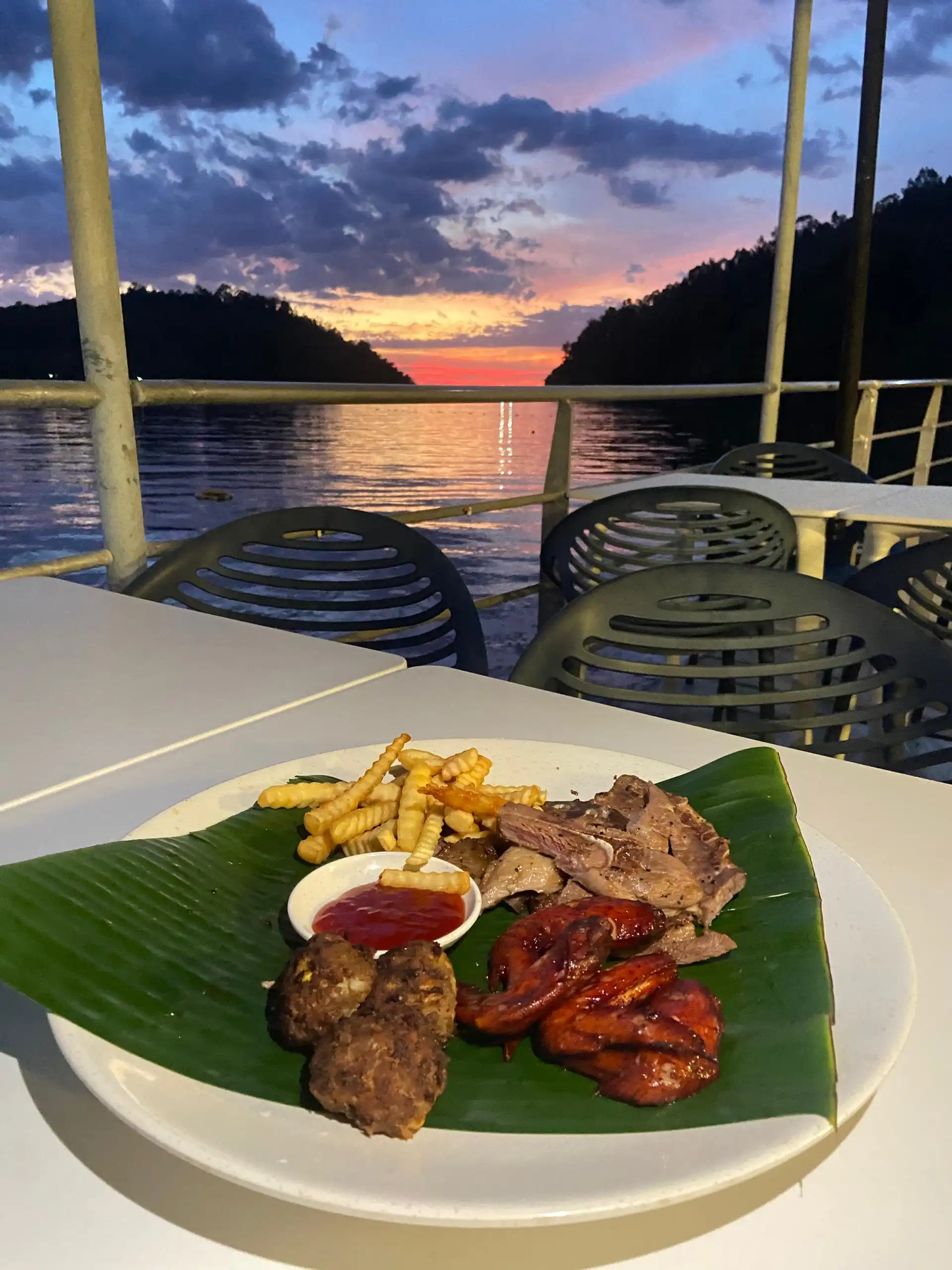 Dinner plate with grilled meats, fries, and sauce on a banana leaf, overlooking a sunset by the water.