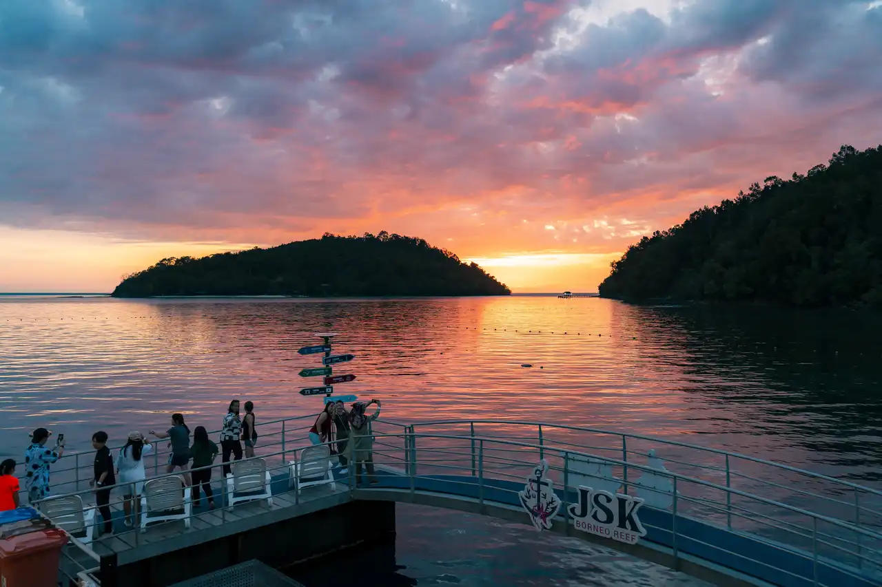 Sunset over a tranquil sea with people on a pier, featuring the JSK Borneo Reef sign.